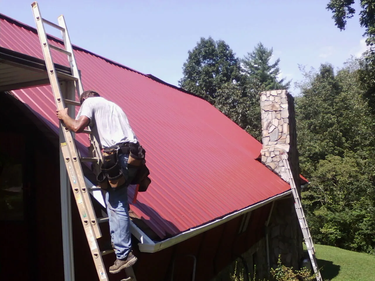 Roofer on a ladder beside a red metal roof and stone chimney — Soffit & Fascia Repair by Roofing Brothers in Rineyville